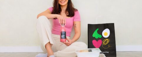 Uśmiechnięta kobieta siedząca na podłodze obok papierowej torby LOVE Catering i zestawu pudełek, trzymająca w dłoni butelkę różowego soku smoothie.