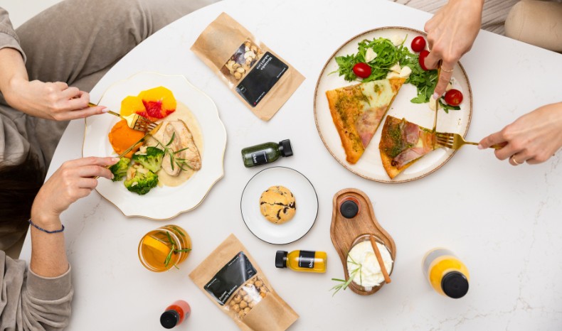 Widok z góry na stół podczas wspólnego posiłku – na talerzach widoczne mięso z brokułami oraz kawałki pizzy, obok prozdrowotne shoty i przekąski w opakowaniach LOVE Catering