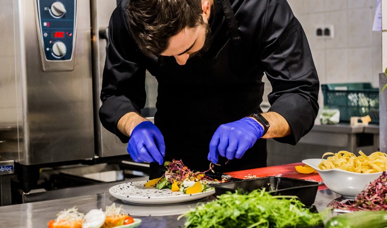 Profesjonalny kucharz w czarnym kitlu i niebieskich rękawiczkach precyzyjnie dekoruje danie na talerzu w nowoczesnej kuchni cateringowej.