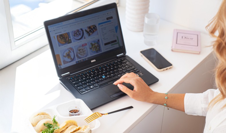 Kobieta pracująca przy laptopie, na którym widoczna jest strona internetowa z ofertą diet, obok komputera leży pudełko z pierogami od cateringu dietetycznego.