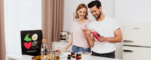 Uśmiechnięta para w jasnej kuchni przygotowująca się do posiłku; mężczyzna otwiera butelkę soku smoothie, obok na blacie leżą pudełka z jedzeniem i torba LOVE Catering.