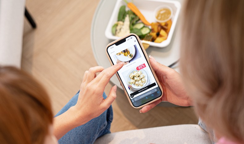 Widok z perspektywy osoby trzymającej smartfon i przeglądającej ofertę diet pudełkowych na stronie internetowej; w tle na stoliku widoczne pudełko z sałatką.