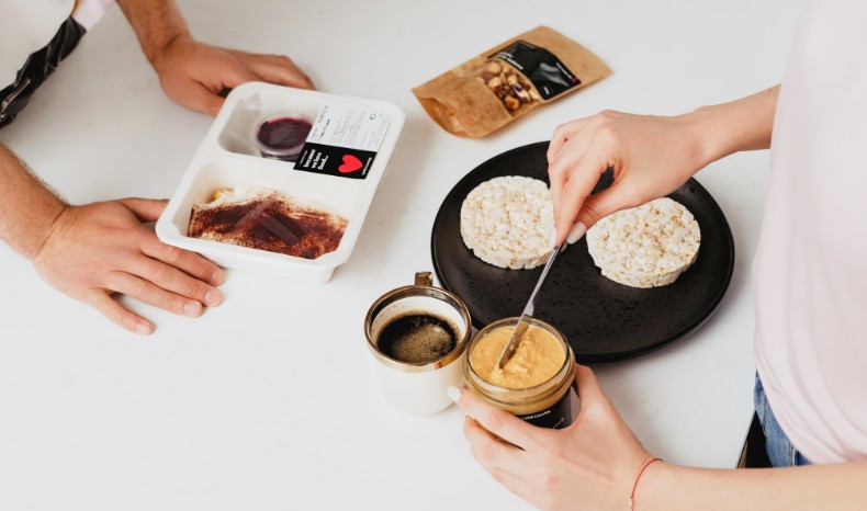Zbliżenie na dłonie smarujące wafle ryżowe masłem orzechowym ze słoika; obok filiżanka czarnej kawy i zamknięty deser w pudełku od Love Catering.