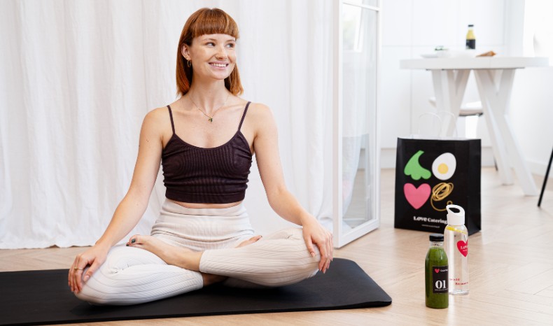 Uśmiechnięta kobieta siedząca w pozycji jogi na macie do ćwiczeń; obok niej butelka zielonego soku i torba papierowa z logo cateringu.
