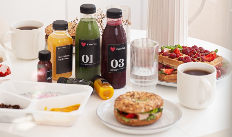 Estetyczne śniadanie na białym stole: butelki z kolorowymi sokami smoothie (01, 03), gofry z malinami, bajgiel z warzywami oraz kawa w białym kubku.