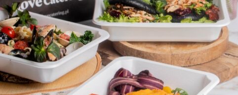 Trzy różne dania cateringu dietetycznego w białych pojemnikach: makaron z owocami morza i pomidorkami, czarne bułeczki bao z nadzieniem oraz pieczona papryka z hummusem i fioletową marchwią.