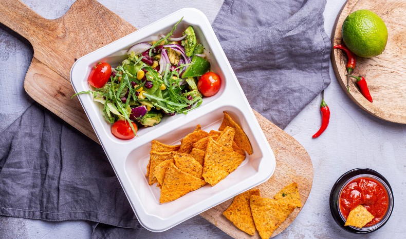 Zdjęcie z góry przedstawiające biały, dwukomorowy pojemnik z cateringiem dietetycznym. W jednej komorze kolorowa sałatka z rukolą, pomidorkami i czerwoną cebulą, w drugiej chrupiące nachosy. Obok miseczka z salsą i świeża limonka.